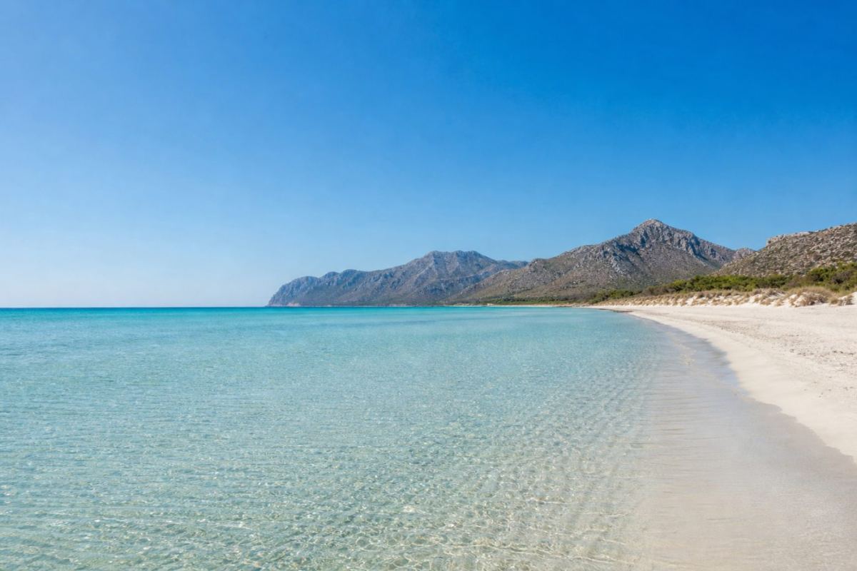 Lång ljus sandstrand med grunt turkost vatten och bergssiluett på norra Mallorca